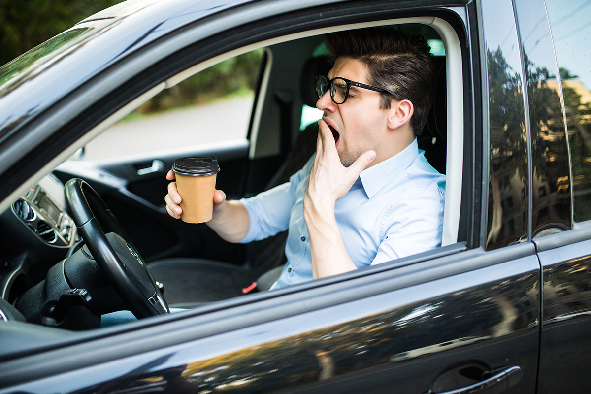 Tired driver behind wheel of car yawning while holding a cup of coffee